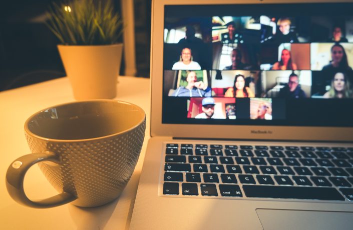 Photograph of a laptop screen showing multiple people on a video call, next to the laptop is a mug
